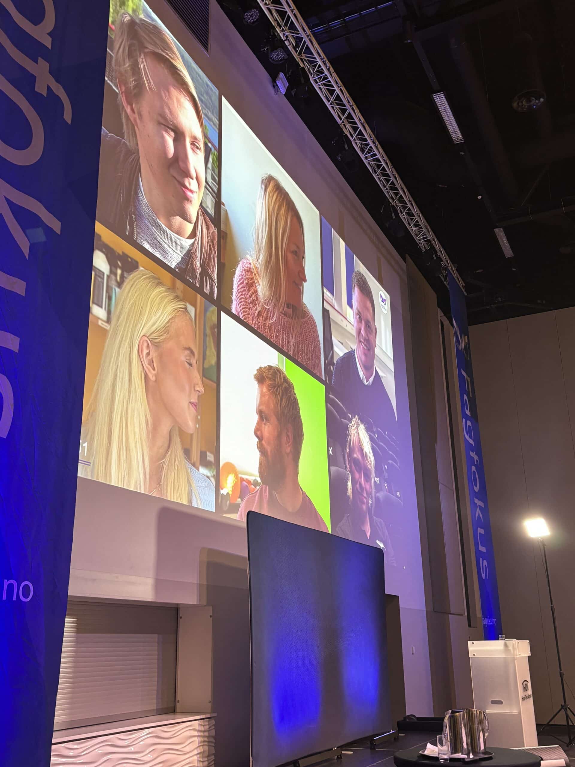 En stor skjerm viser en videosamtale med seks personer i et konferanserom, med blå bannere og scenelys synlig.