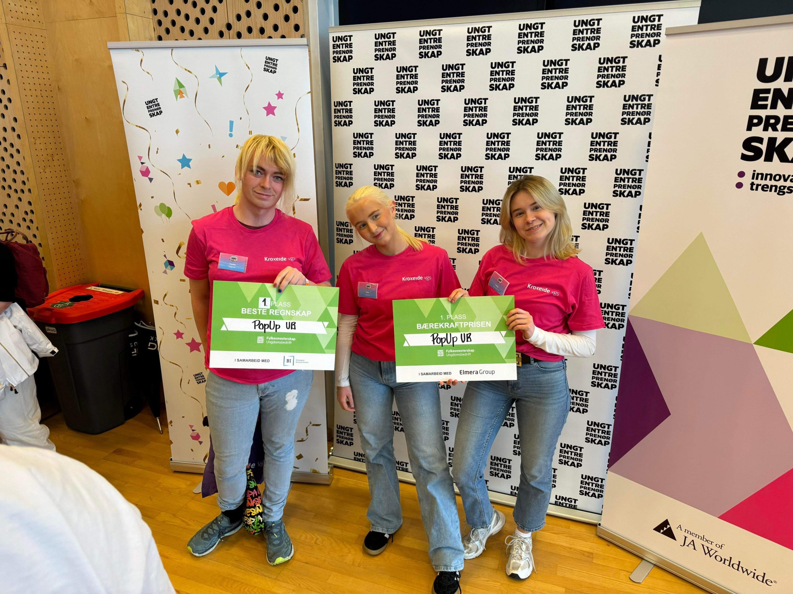 Tre personer i rosa skjorter med grønne sertifikater står innendørs på et arrangement med bannere i bakgrunnen.