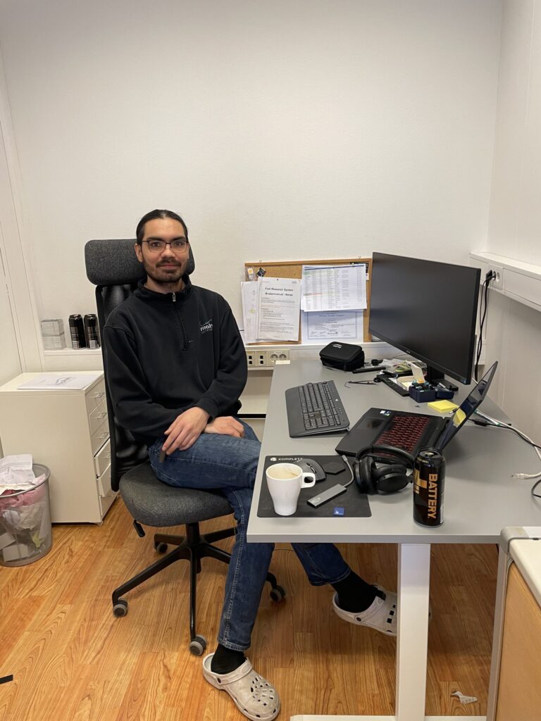 En person sitter ved et skrivebord med datamaskin, tastatur, hodetelefoner, kaffekopp og energidrikk på et kontor med hvite vegger og tregulv.