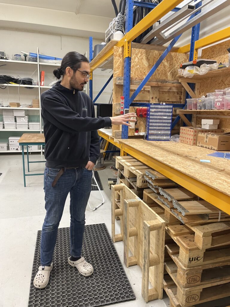 En mann med langt hår i hestehale, briller, mørk genser, jeans og hvite tresko, står i et verksted og peker på plastskuffer på en lagerhylle.