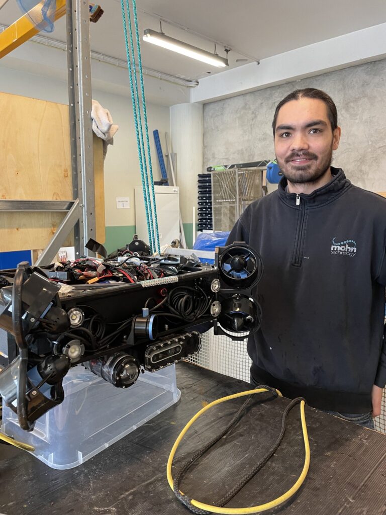 En person står ved siden av en kompleks robot med synlige ledninger og flere propeller i et verksted.