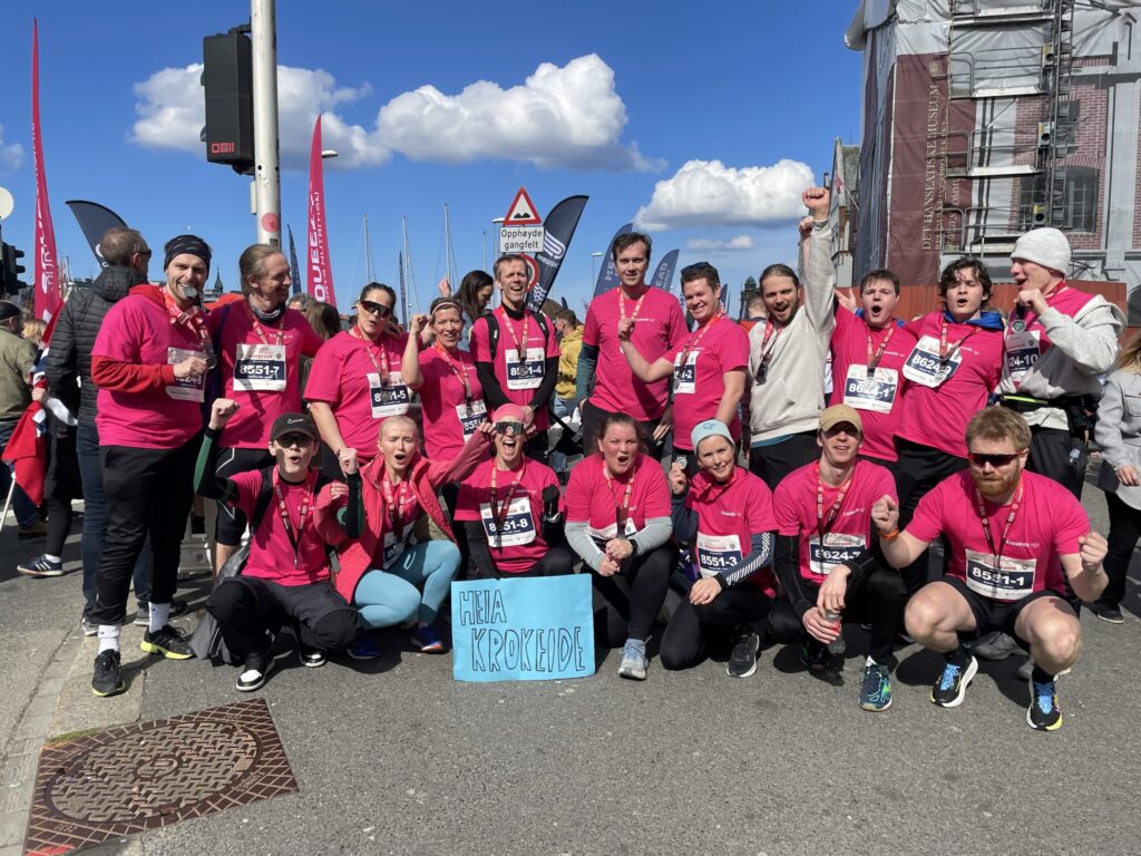 IMG_1769-scaled En gruppe løpere i matchende rosa skjorter poserer sammen utendørs etter et løp, med medaljer og et blått skilt med påskriften "Hela Krockede".