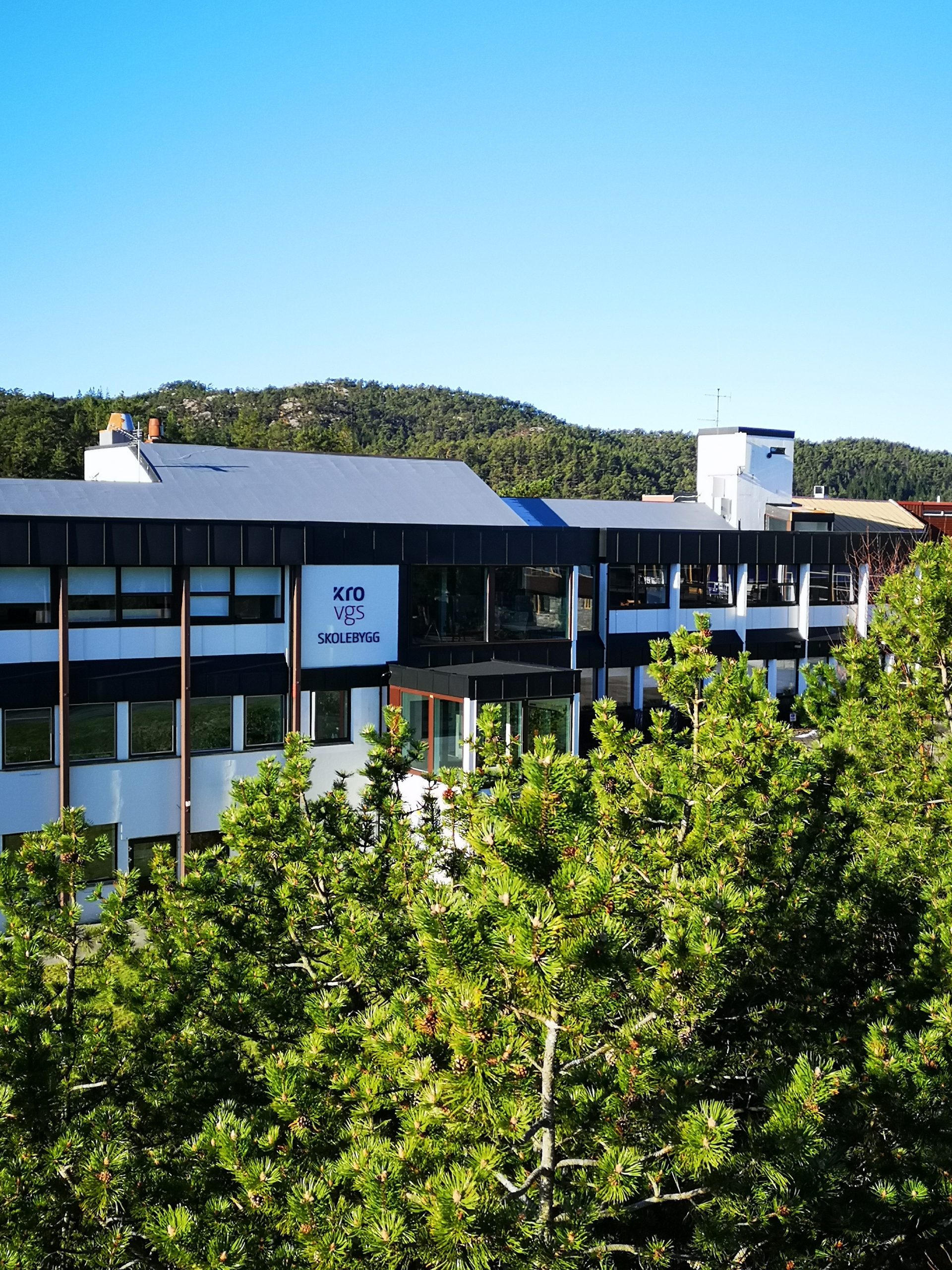 En moderne bygning i flere etasjer med skiltet "KRO VGS SKOLEBYGG" er delvis skjult av furutrær, med en skogkledd ås og klarblå himmel i bakgrunnen.