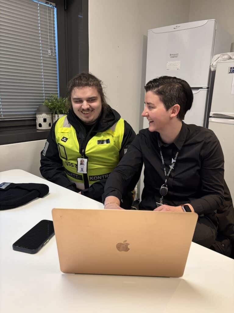 To personer sitter ved et bord, den ene i sikkerhetsuniform og den andre i svart antrekk, og begge ser på en bærbar datamaskin og smiler i et kontormiljø.