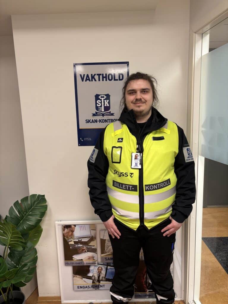 En sikkerhetsvakt i gul vest står innendørs ved siden av et veggskilt med påskriften "Vakthold Skan-Kontroll".