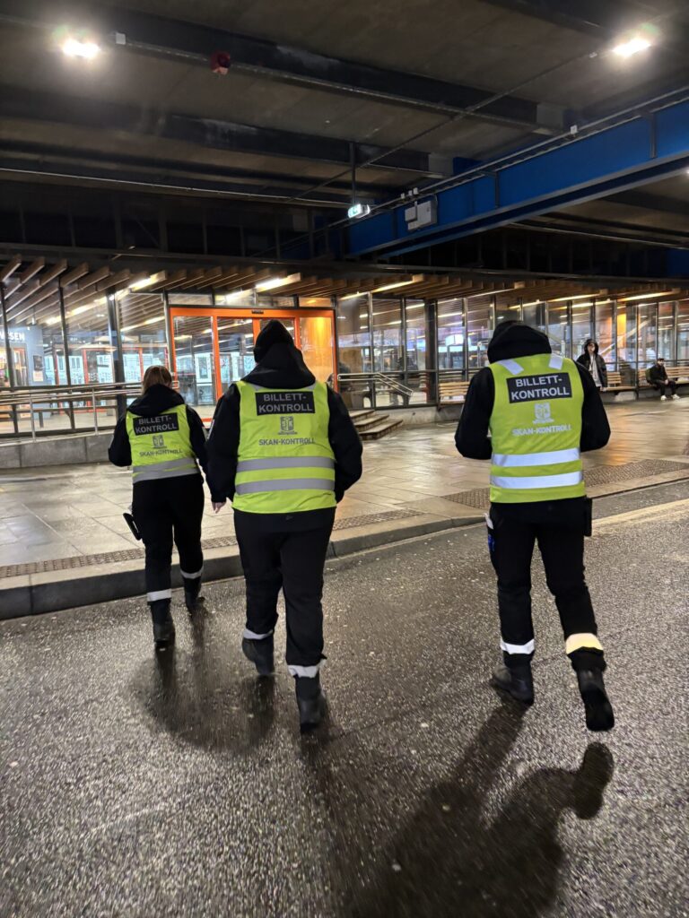 Tre billettkontrollører i refleksvester går i nærheten av en transportterminal med glassvegger om natten.