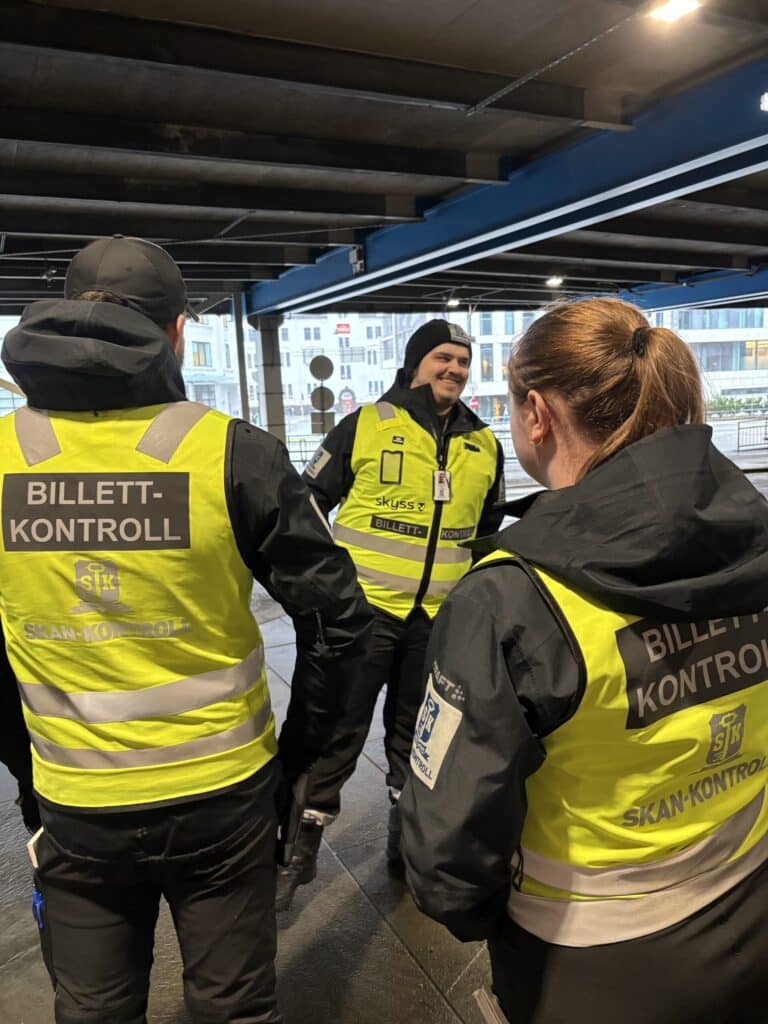 Tre uniformerte kontrollører med gule vester merket "BILLETT-KONTROLL" står sammen innendørs og ser ut til å snakke sammen og kontrollere billetter.