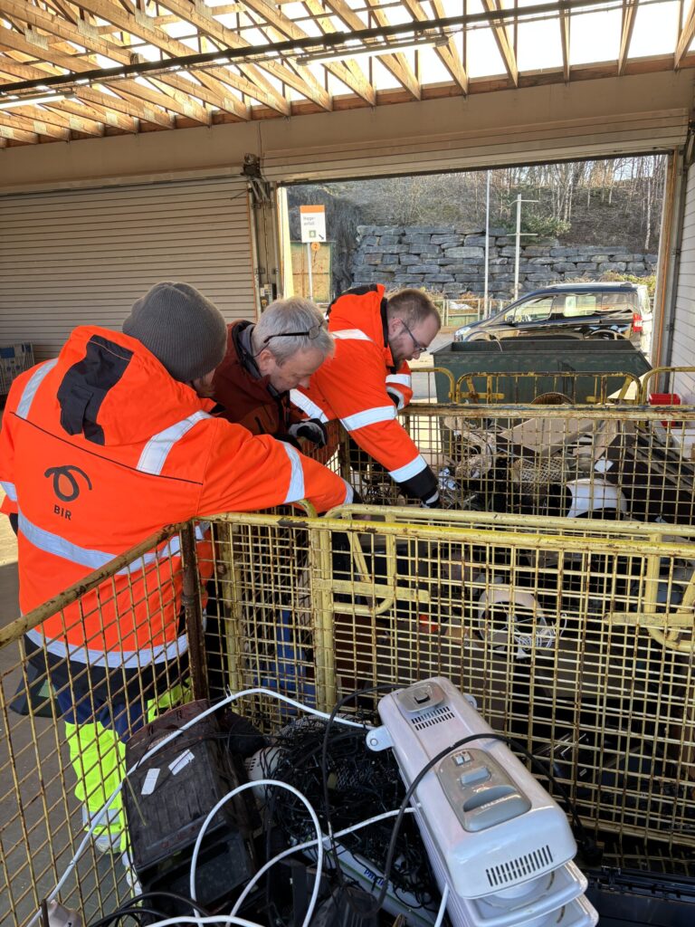 Tre personer iført vernetrøyer undersøker og sorterer elektronisk avfall i en inngjerdet innhegning på et gjenvinningssenter.