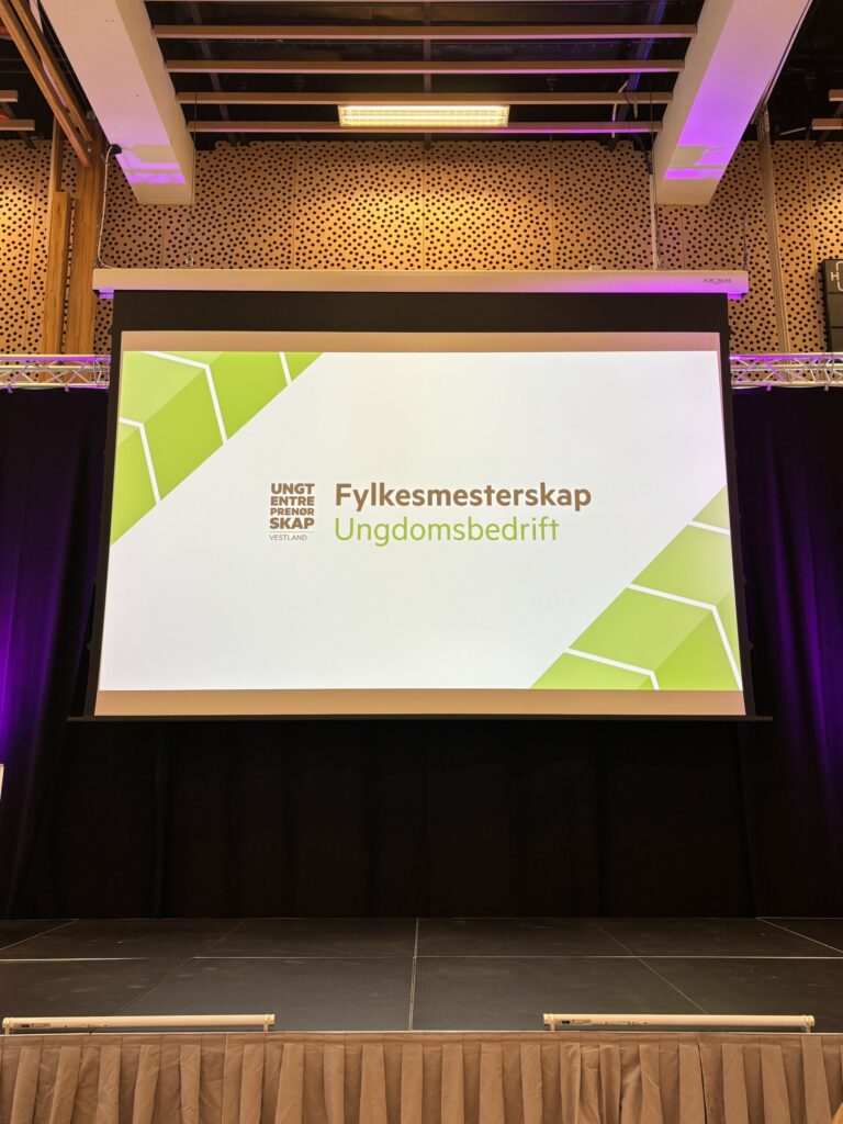 På en projeksjonsskjerm vises "Fylkesmesterskap Ungdomsbedrift" med et grønt geometrisk mønster, på en scene med svarte gardiner og trepanel i bakgrunnen.
