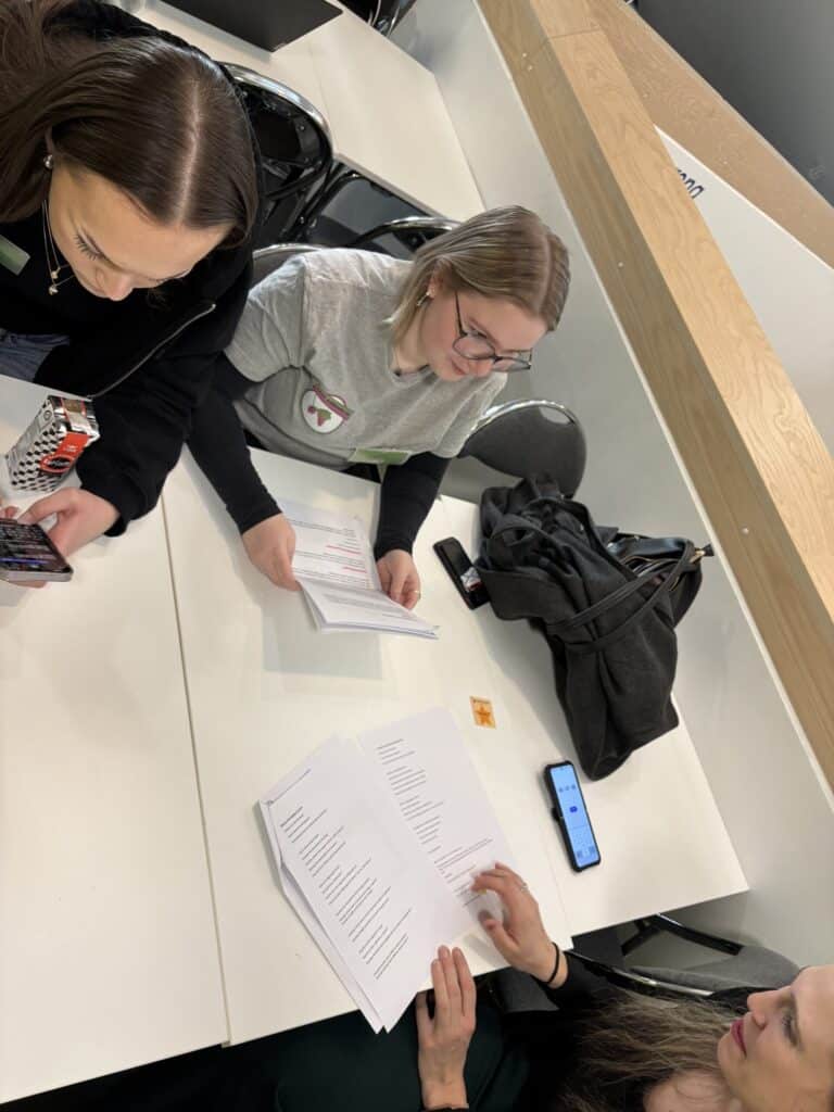 Tre personer sitter ved et bord med papirer, en smarttelefon og en snackskartong og går gjennom dokumenter sammen i et klasserom eller møterom.