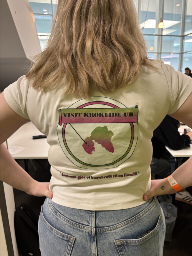 En person står med hendene på hoftene, iført en skjorte med teksten "Visit Krokeide UB" og en fargerik grafikk på ryggen, i et moderne innemiljø.