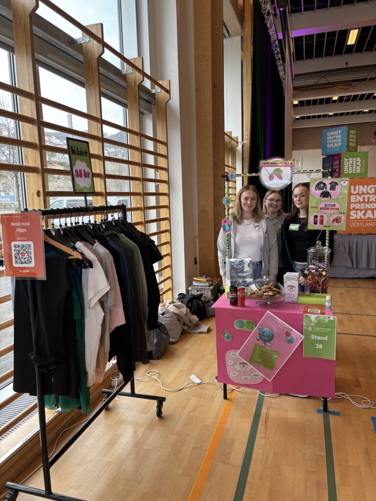 Tre personer står bak en rosa bod med snacks og flyers på et innendørsarrangement. Til venstre står et klesstativ, og bak dem henger informasjonsplakater.
