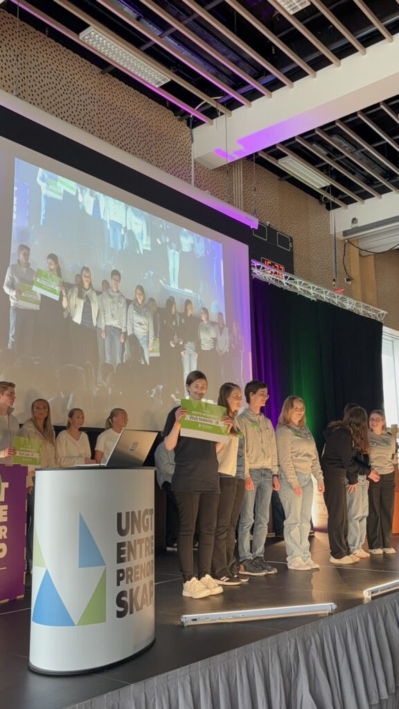 En gruppe ungdommer står på scenen og holder et grønt skilt, mens bildet av dem projiseres på en storskjerm bak dem på et arrangement.