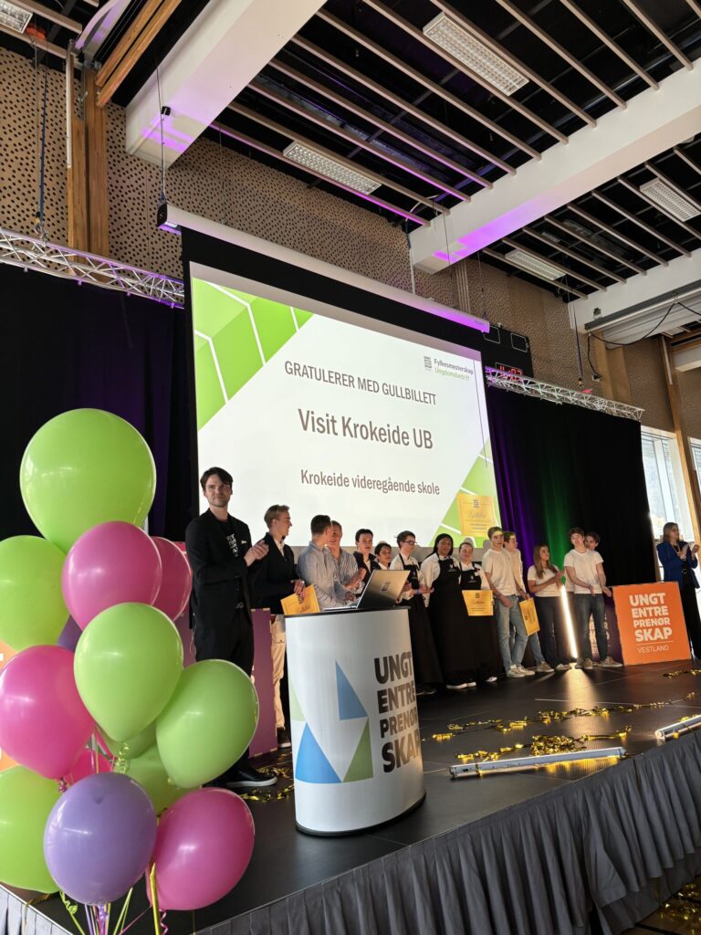 En gruppe mennesker står på scenen på et arrangement med ballonger og en storskjerm bak seg som viser "Visit Krokeide UB" og "GRATULERER MED GULLBILLETT".