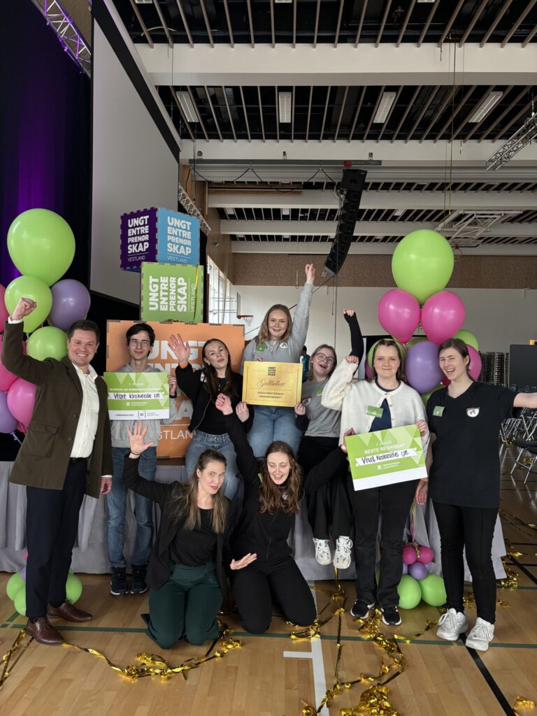 En gruppe ungdommer og en voksen står og kneler innendørs, med diplomer og skilt i hendene, og armene hever seg i feiringen, omgitt av fargerike ballonger og bannere.