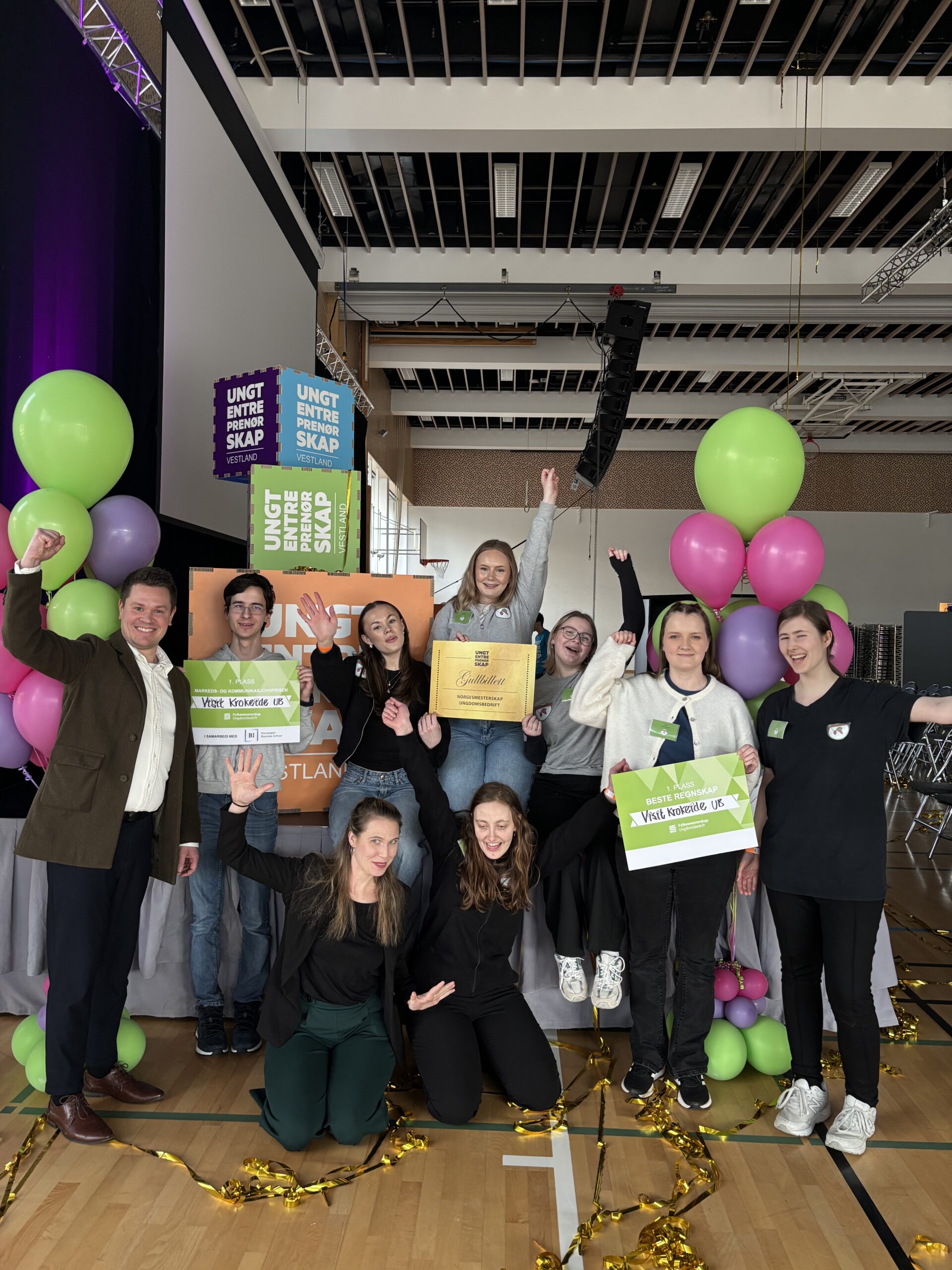 En gruppe mennesker poserer innendørs med diplomer og skilt i hendene, omgitt av ballonger og bannere, for å feire en prestasjon.