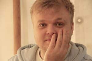 En person med blondt hår og lyst skjegg tar seg til ansiktet med den ene hånden og ser direkte inn i kameraet. Bakgrunnen er svakt opplyst og nøytral.