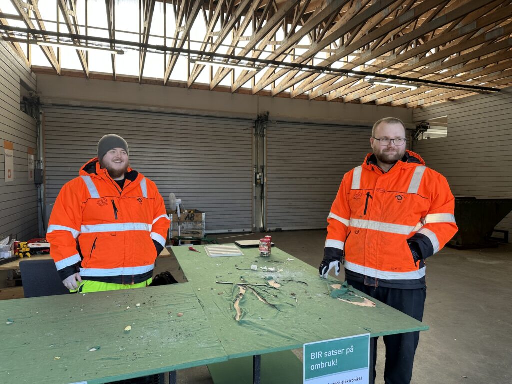 To menn i oransje vernetrøyer står bak et grønt bord med diverse materialer på, inne i en lagerbygning med delvis åpent tak og metallvegger.