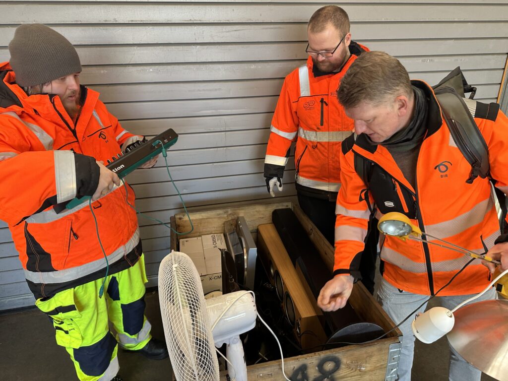 Tre menn i oransje vernetrøyer inspiserer og sorterer en trekasse med diverse brukt elektronikk, deriblant en vifte og en lampe, foran en metallvegg.