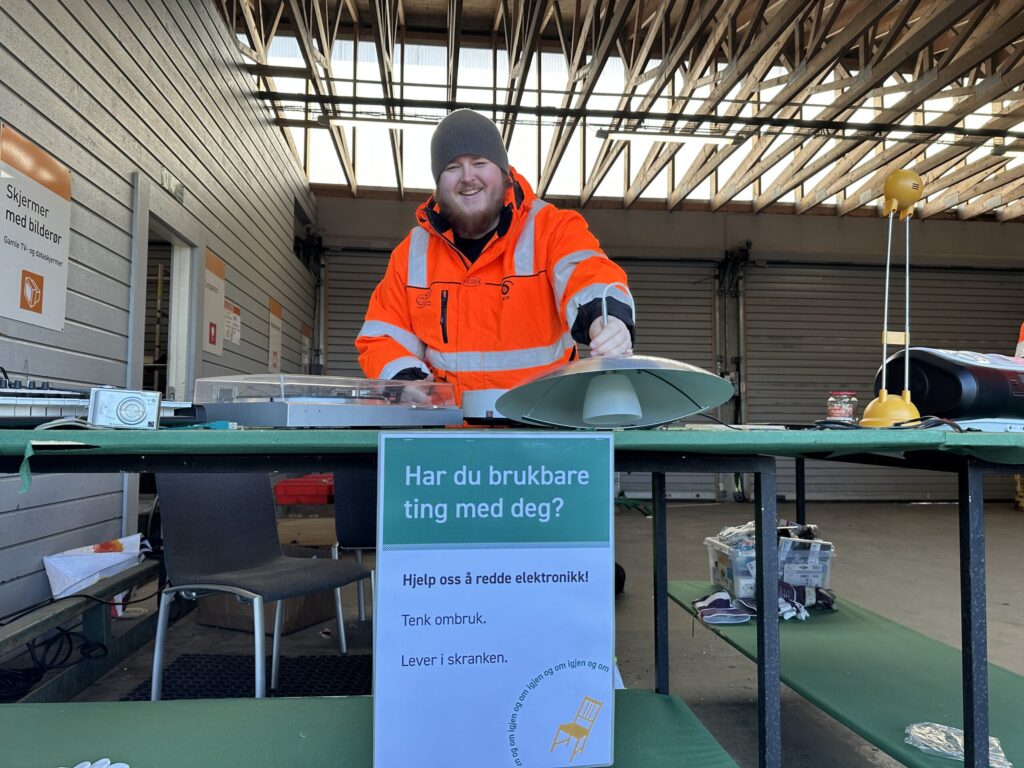 En person i refleksjakke står bak et bord med brukt elektronikk på et gjenbrukssenter. Et skilt på norsk spør om besøkende har gjenbrukbare gjenstander.