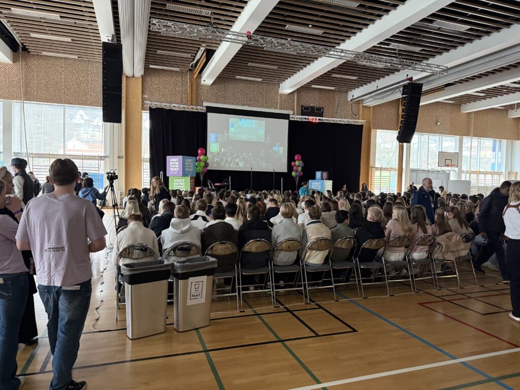 IMG_6251 En stor gruppe mennesker sitter på rekke og rad foran en scene med skjerm og ballonger i en gymsal under et innendørs arrangement eller en samling.