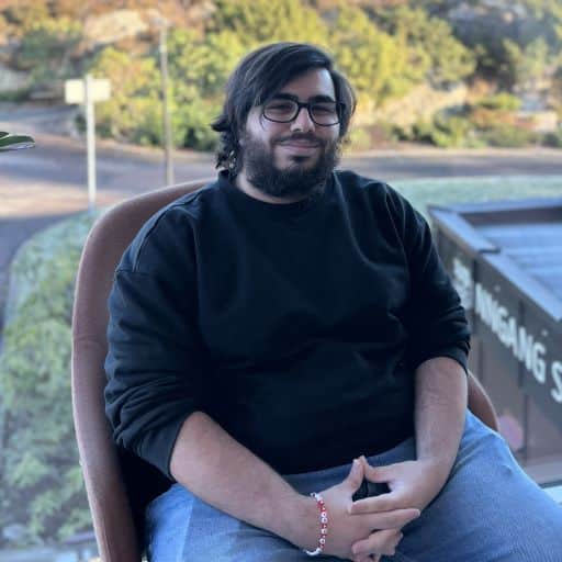 En person med langt mørkt hår, skjegg og briller sitter utendørs på en stol, iført svart genser og jeans, med et avslappet uttrykk.