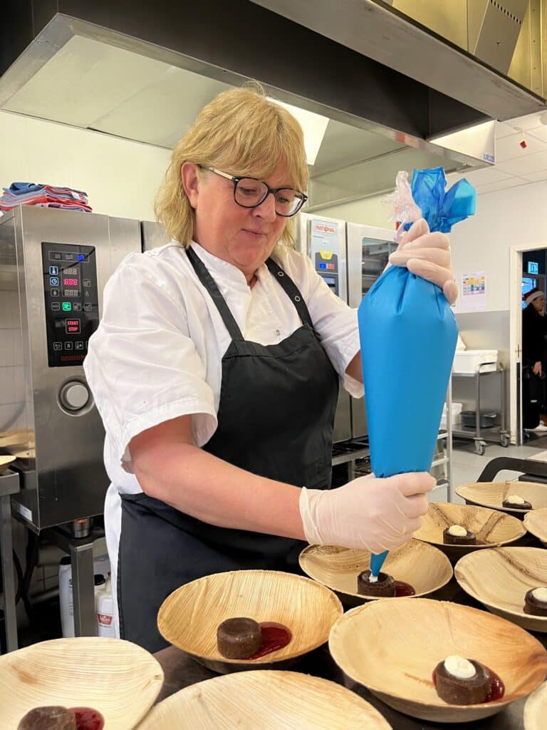 En kokk i hvit uniform og svart forkle bruker en blå sprøytpose til å fylle små, runde kaker i treboller med krem.