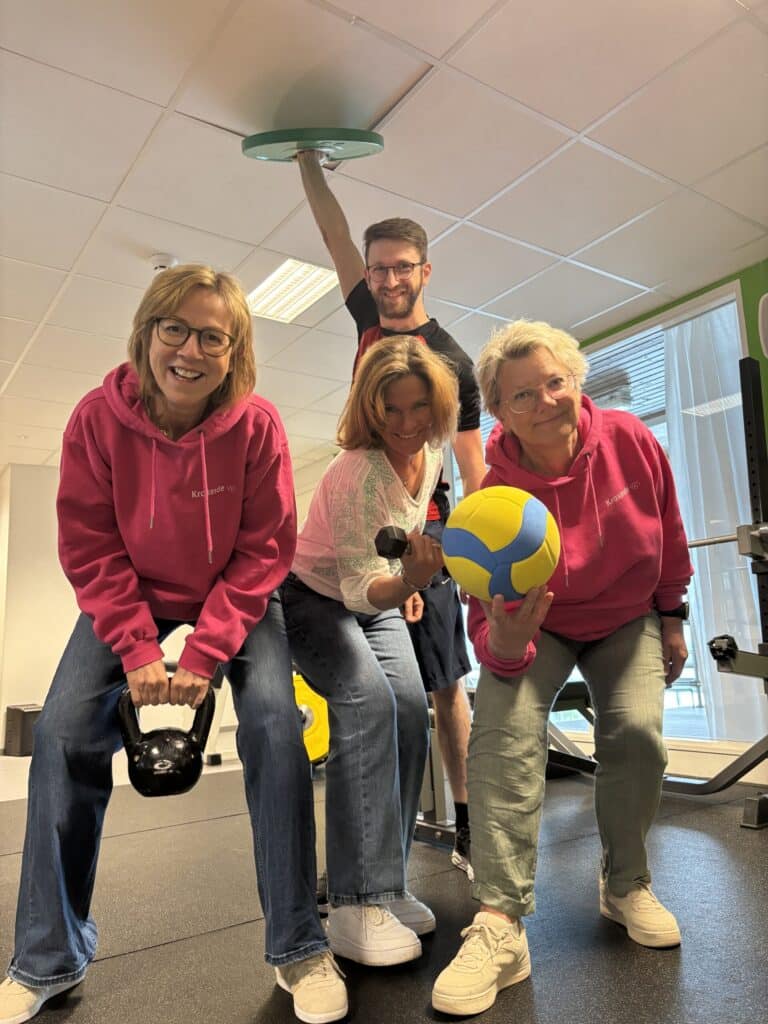 Fire voksne i et treningsstudio, tre kvinner i rosa hettegensere som holder i treningsapparater, og en mann som står bak dem og holder seg fast i en takarmatur.