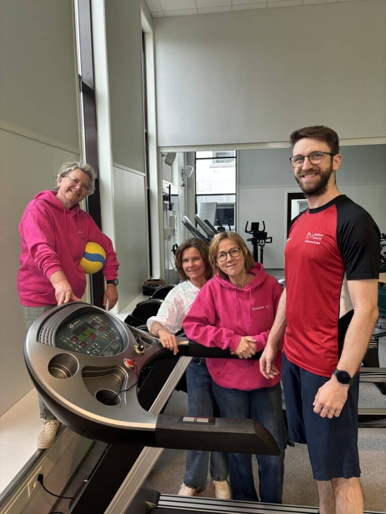 Fire voksne står rundt en tredemølle i et treningssenter; tre kvinner har rosa hettegensere, og en mann har på seg rød sportsskjorte og shorts.