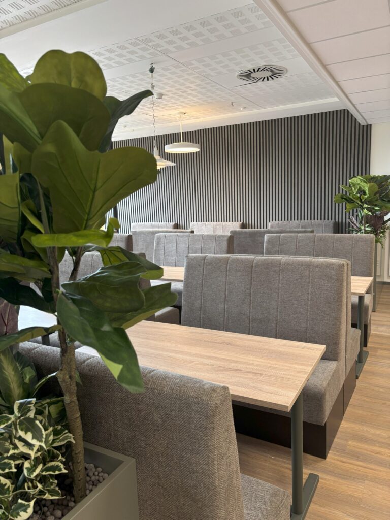 Kantine-13082025-3-scaled Moderne kafeteria med grå, polstrede sitteplasser, lyse trebord og flere store potteplanter; vertikal lamellvegg i bakgrunnen.