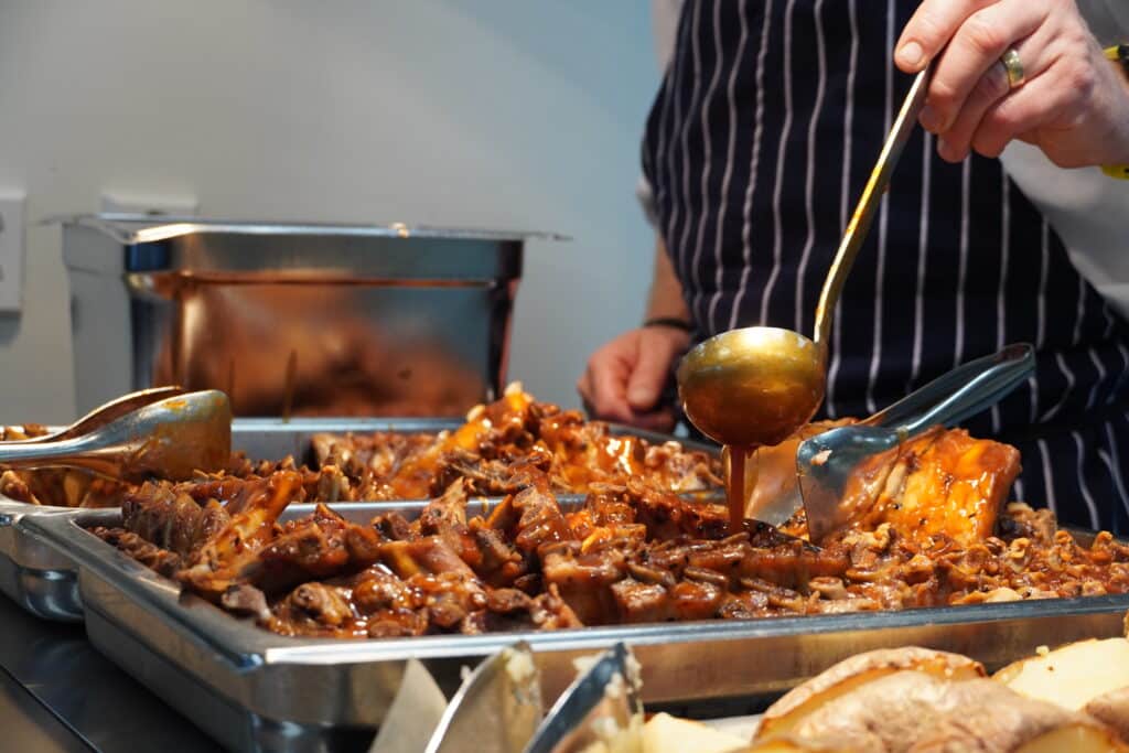 En person i et stripete forkle heller saus fra en øse over et metallfat med svineribbe.