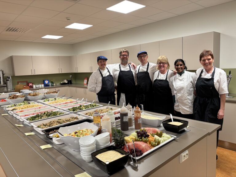 Seks kjøkkenmedarbeidere i uniformer står bak en disk fylt med brett med diverse mat på et storkjøkken.