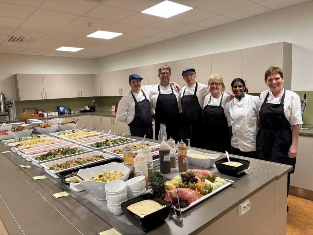 Kjokkengjengen-scaled Seks kjøkkenmedarbeidere i uniformer og forklær står bak en lang disk fylt med brett med tilberedt mat på et storkjøkken.