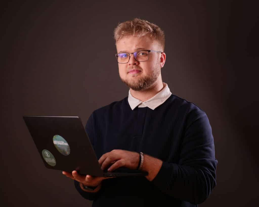 Kristian_UiB En mann med briller og skjegg holder og skriver på en bærbar datamaskin foran en vanlig, mørk bakgrunn.