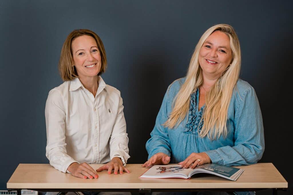 To kvinner sitter ved et bord mot en mørk bakgrunn, den ene i hvit skjorte, den andre i blå bluse, med et oppslått magasin foran seg.
