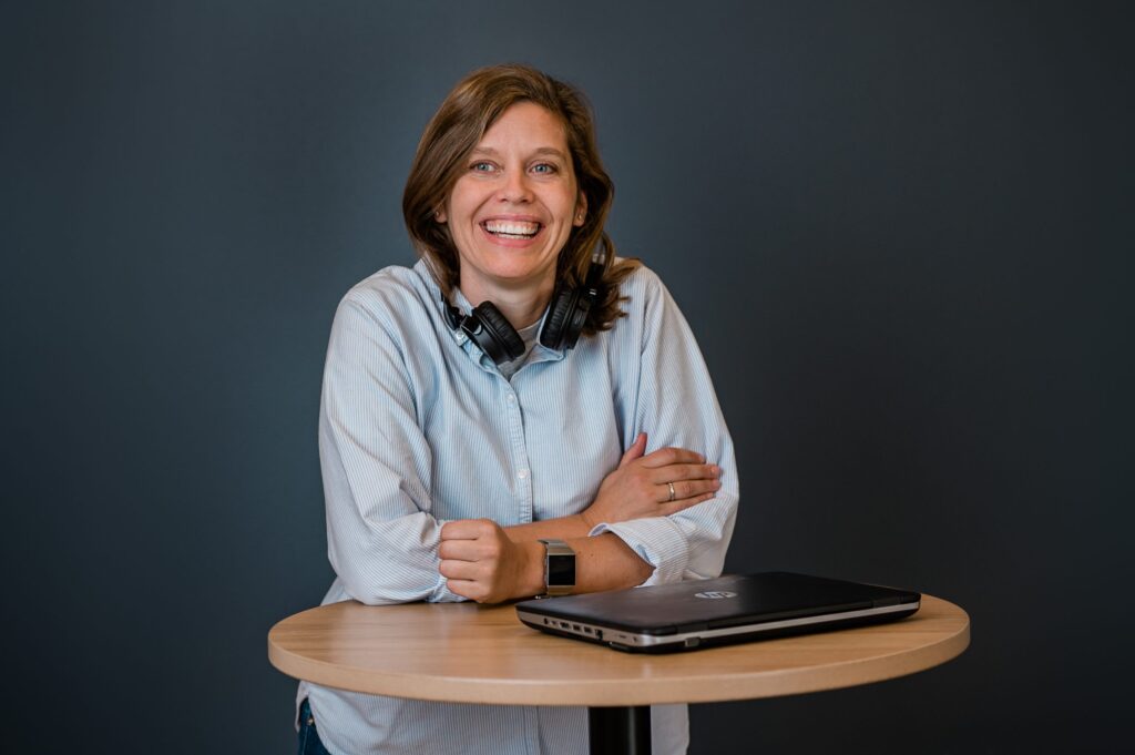 Krokeide2021-26-scaled En person med lys skjorte og hodetelefoner rundt halsen smiler mens han sitter ved et rundt bord med en lukket bærbar PC.