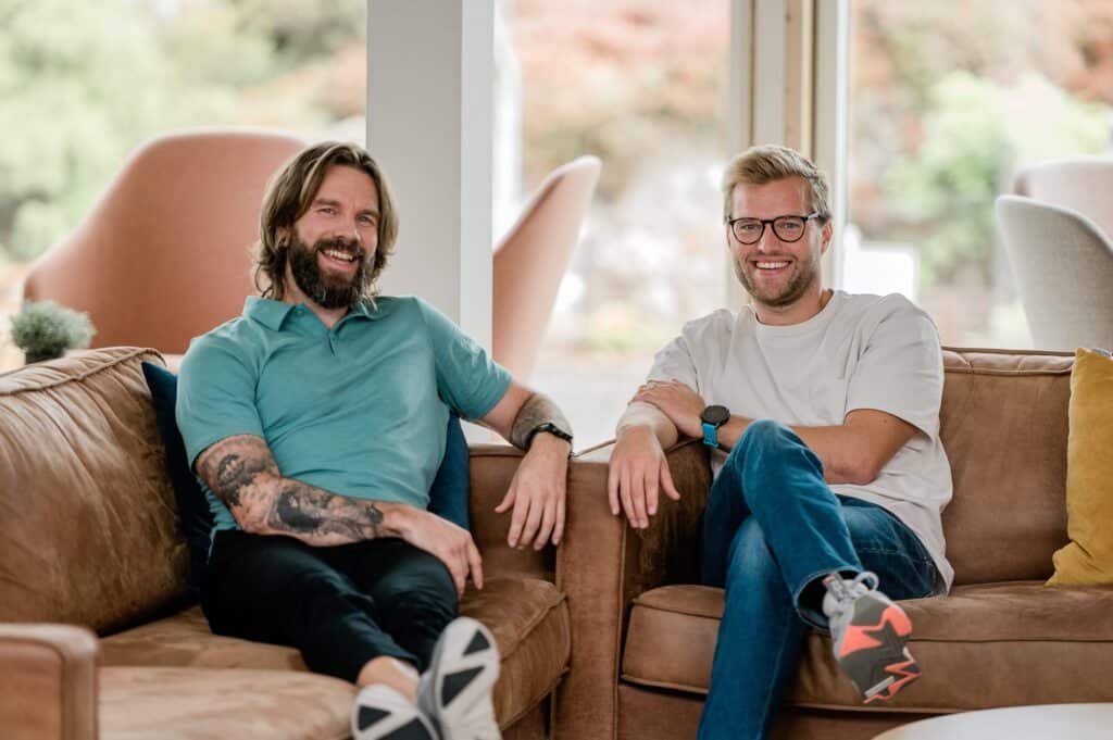 To menn sitter i en brun sofa, smilende og uformelt kledd, med naturlig lys inn gjennom vinduer i bakgrunnen.