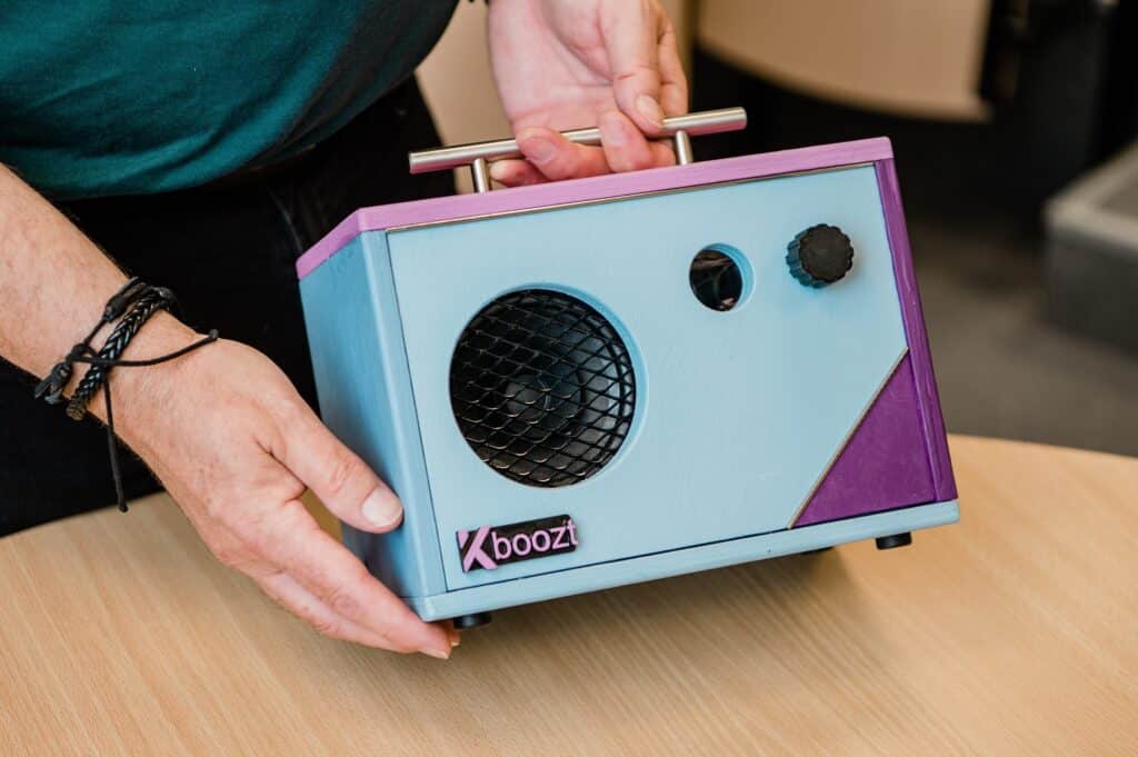 Krokeide2021-55-scaled Person som holder en blå og lilla rektangulær høyttaler med et håndtak, merket "Xboozl", plassert på en lys treflate.