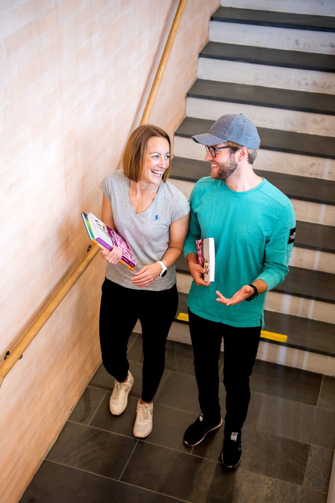 To personer holder bøker og snakker mens de går opp en trapp innendørs.