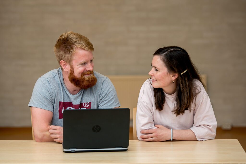 To personer sitter ved et bord vendt mot hverandre, med en bærbar datamaskin foran seg. De ser ut til å ha en samtale, og begge smiler.
