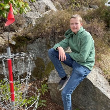 En ung person i grønn hettegenser og jeans sitter på en stein ved siden av en discgolfkurv utendørs.