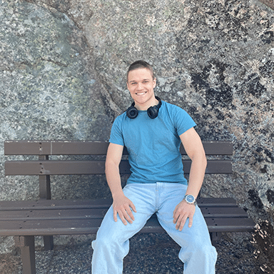 En ung mann i blå t-skjorte og jeans sitter på en brun benk utendørs med hodetelefoner rundt halsen og smiler til kameraet. Bakgrunnen er en steinmur.