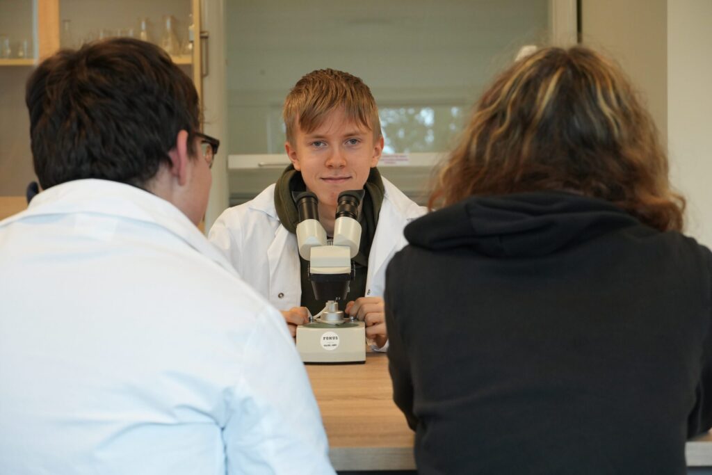 En ung person i laboratoriefrakk sitter ved et bord med et mikroskop, vendt mot to andre i samtale.