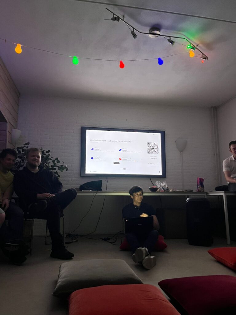 En gruppe mennesker sitter og står i et svakt opplyst rom med en presentasjon på en stor skjerm og fargerike lyskæder over hodet.