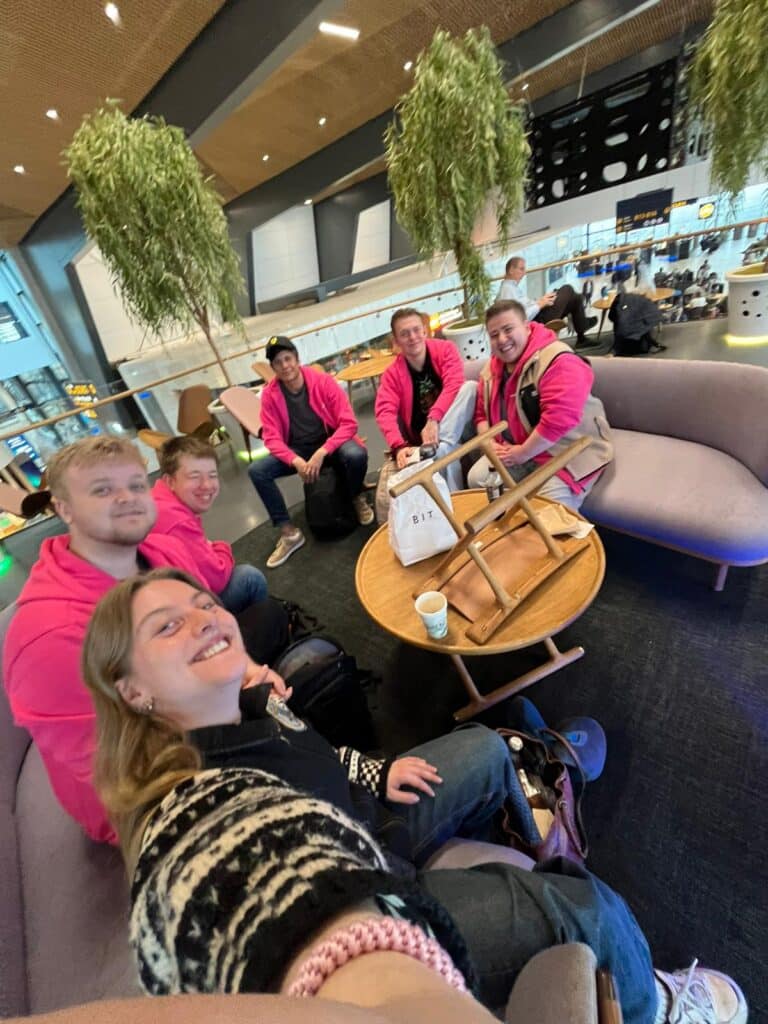 Seks personer sitter rundt et bord i et moderne innelokale. Fem har matchende rosa hettegensere, og én tar en gruppeselfie. På bordet står det drikke og vesker.
