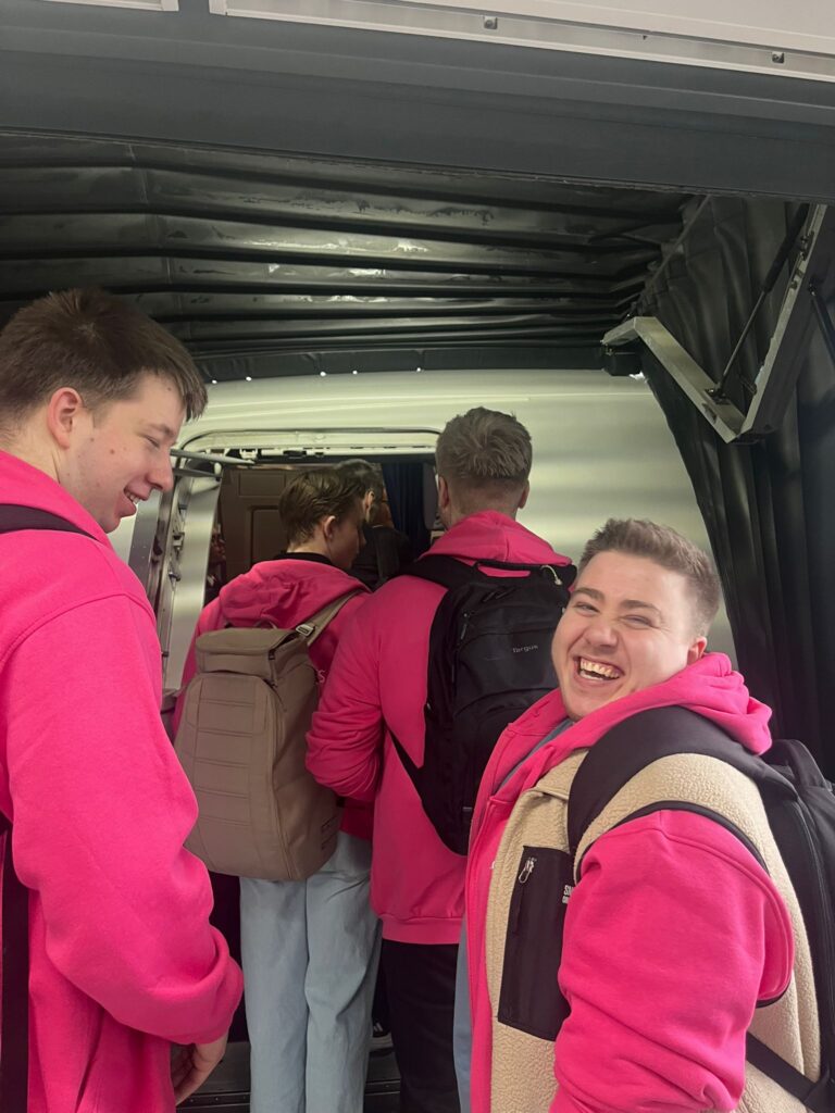 Fire personer iført rosa hettegensere og ryggsekker går om bord i et fly gjennom en jetbro; én person smiler til kameraet.