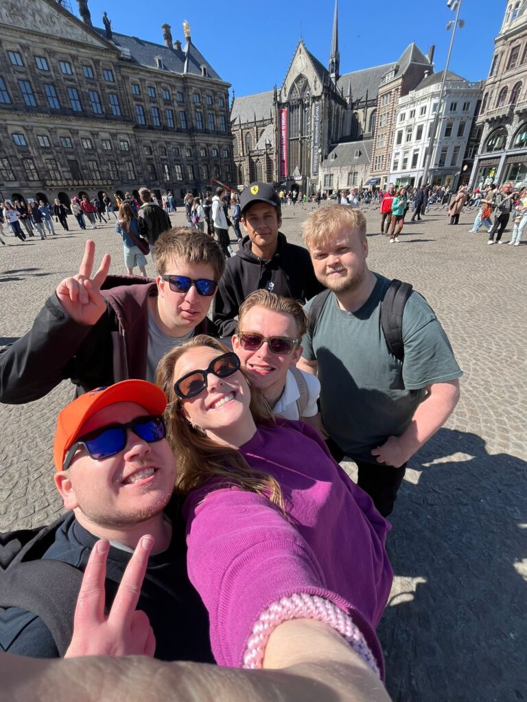 En gruppe på seks personer tar en selfie på et travelt europeisk torg med historiske bygninger og en kirke i bakgrunnen på en solrik dag.
