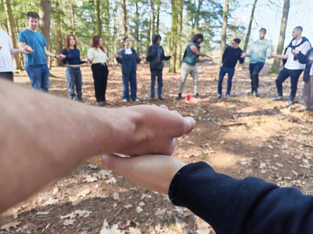 En gruppe mennesker står i en sirkel i en skog og holder hverandre i hendene. I forgrunnen ser man to hender som er knyttet sammen, mens gruppen er litt ute av fokus i bakgrunnen.