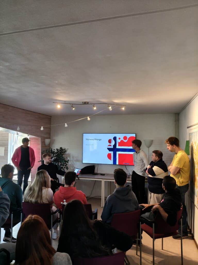 En gruppe mennesker sitter og lytter til en presentasjon om Norge, med en stor TV-skjerm som viser det norske flagget og informasjon.