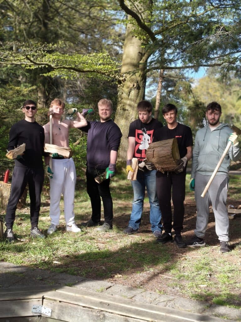 Seks unge menn står utendørs i et skogsområde med tømmerstokker, ved og verktøy i hendene, lettkledde, med trær og sollys i bakgrunnen.